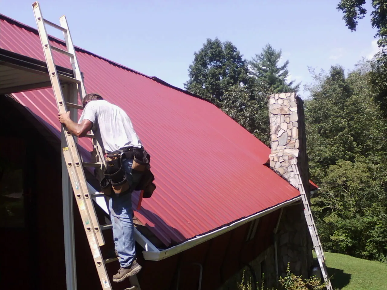 Roofer on a ladder beside a red metal roof and stone chimney — Soffit & Fascia Repair by Roofing Brothers in Baldwin